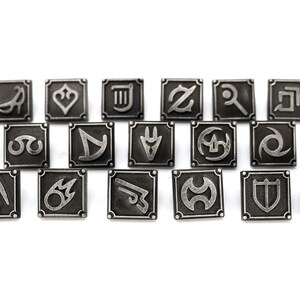 May include: A set of 18 silver-toned metal pins featuring various symbols, including a heart, a musical note, a sword, and a shield. The pins are arranged in rows on a white background.