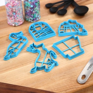 Op de afbeelding: Een set blauwe plastic koekjesvormen in de vorm van een vogel, een kat, een huis, een schommel en een raam. De vormen liggen op een houten snijplank met een blauwe spatel en twee potten met sprinkles.