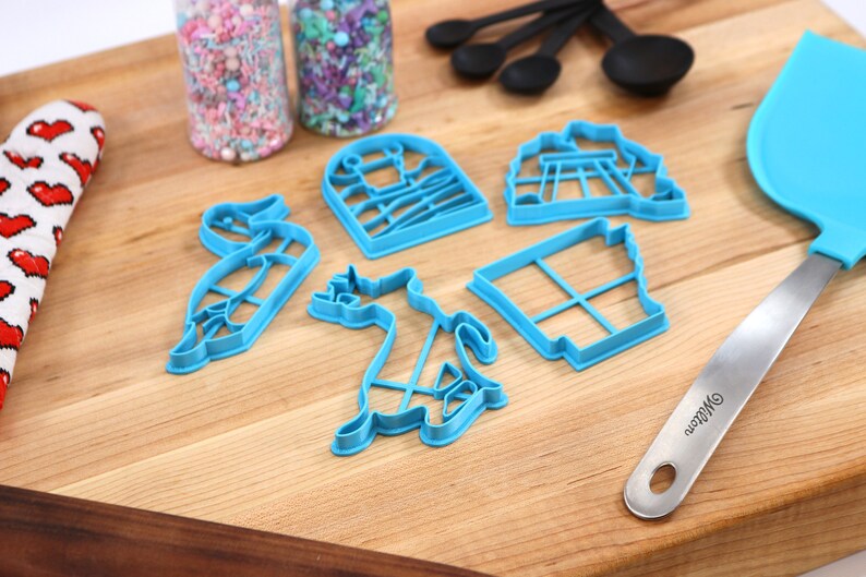 Op de afbeelding: Een set blauwe plastic koekjesvormen in de vorm van een vogel, een kat, een huis, een schommel en een raam. De vormen liggen op een houten snijplank met een blauwe spatel en twee potten met sprinkles.