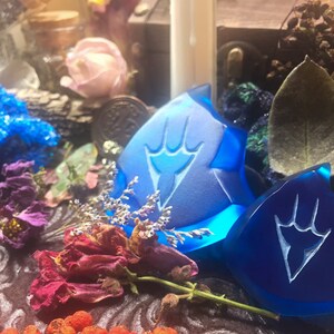 May include: Two blue glass charms with a white design of a hand with three fingers. The charms are on a dark brown surface with dried flowers and other decorative items.