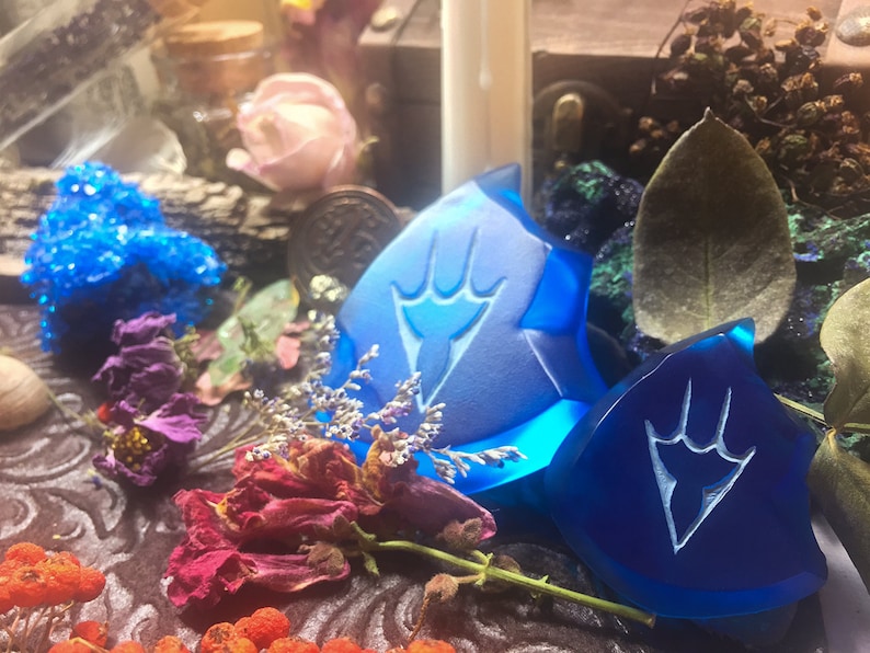 May include: Two blue glass charms with a white design of a hand with three fingers. The charms are on a dark brown surface with dried flowers and other decorative items.