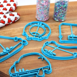 May include: A set of six blue plastic cookie cutters shaped like a car, a house, a hat, a pair of roller skates, a swing set, and a donut.