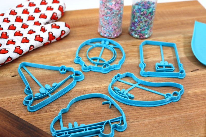 May include: A set of six blue plastic cookie cutters shaped like a car, a house, a hat, a pair of roller skates, a swing set, and a donut.
