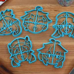 May include: Five blue plastic cookie cutters shaped like cartoon faces. The cutters are on a wooden surface.