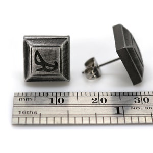 May include: A pair of silver square stud earrings with a black design. The earrings are approximately 1 cm in size.