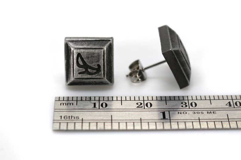 May include: A pair of silver square stud earrings with a black design. The earrings are approximately 1 cm in size.
