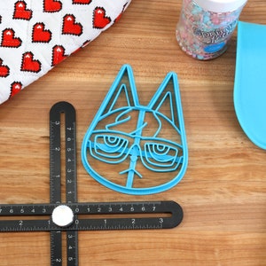May include: A blue plastic cookie cutter shaped like a cat wearing glasses. The cutter is on a wooden surface with a ruler and a blue spatula.