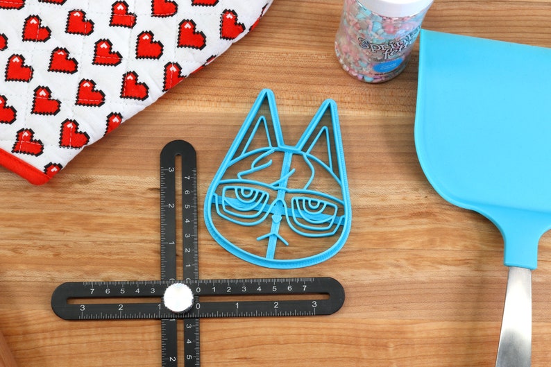 May include: A blue plastic cookie cutter shaped like a cat wearing glasses. The cutter is on a wooden surface with a ruler and a blue spatula.