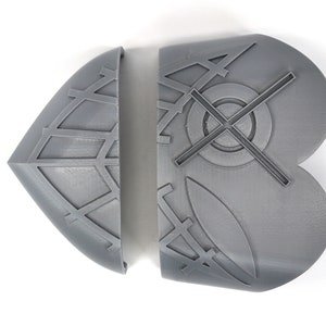 May include: Two-piece, gray, geometric-patterned object. Each piece has a curved edge and a flat back. The left piece is diamond-shaped with a grid pattern. The right piece is a rounded shape with a cross and circle design.