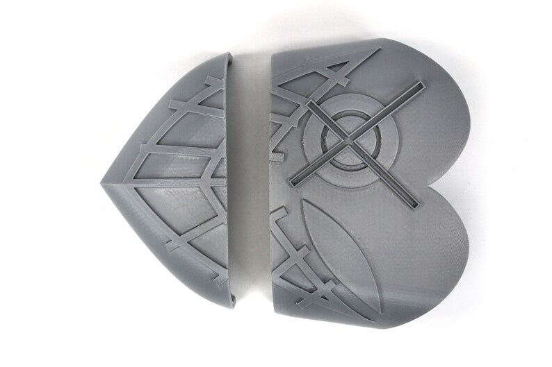May include: Two-piece, gray, geometric-patterned object. Each piece has a curved edge and a flat back. The left piece is diamond-shaped with a grid pattern. The right piece is a rounded shape with a cross and circle design.