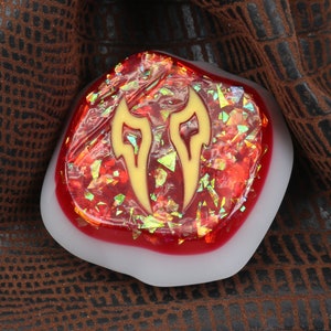 May include: A red and gold resin cabochon with a stylized flame design. The cabochon is set in a white resin base.