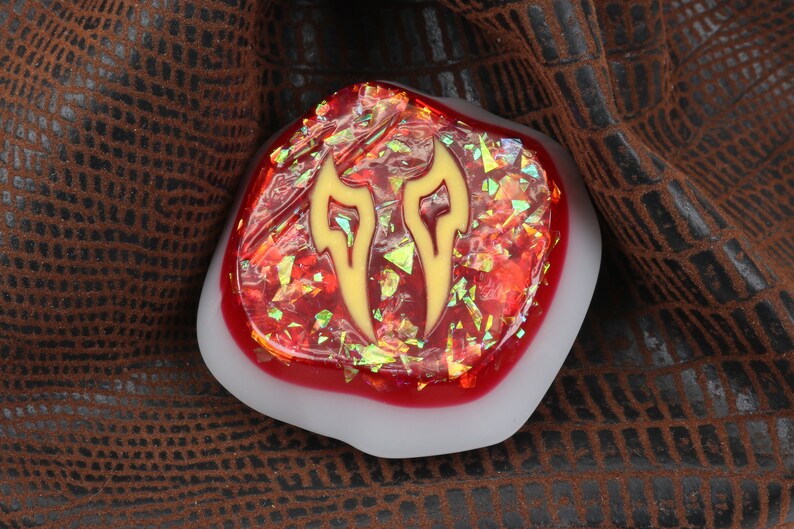 May include: A red and gold resin cabochon with a stylized flame design. The cabochon is set in a white resin base.