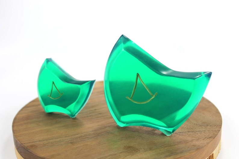 May include: Two green glass paperweights with a gold sailboat design. The paperweights are on a wooden surface.