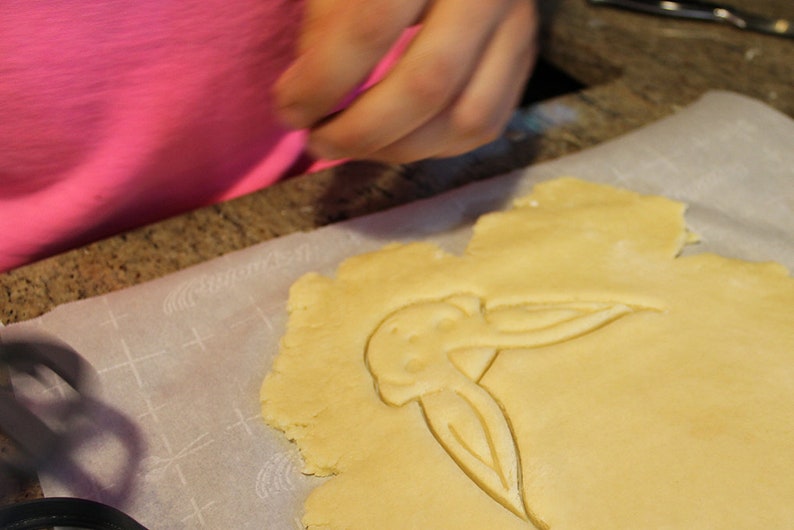 May include: A close-up of a cookie dough cutout in the shape of a bird on a white baking sheet lined with parchment paper.