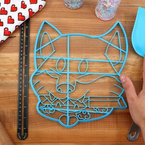 May include: Blue plastic cookie cutter shaped like a cartoon fox with a detailed design. The cutter is on a wooden surface.
