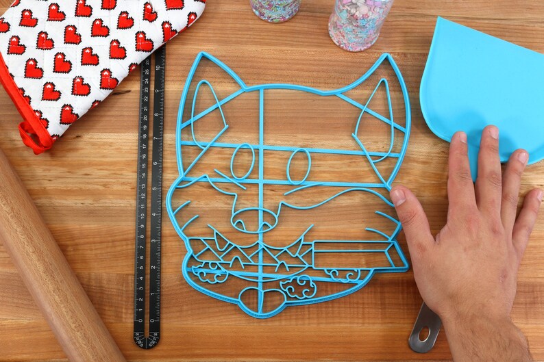 May include: Blue plastic cookie cutter shaped like a cartoon fox with a detailed design. The cutter is on a wooden surface.