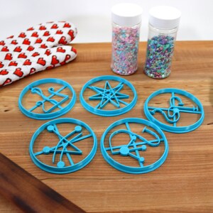May include: Five blue plastic cookie cutters shaped like different geometric designs, including a star, a circle with lines, and a circle with a curved line. The cutters are on a wooden surface with a rolling pin, a spatula, and two jars of sprinkles.
