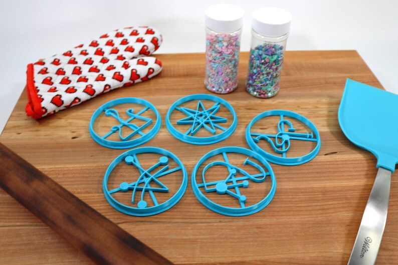 May include: Five blue plastic cookie cutters shaped like different geometric designs, including a star, a circle with lines, and a circle with a curved line. The cutters are on a wooden surface with a rolling pin, a spatula, and two jars of sprinkles.