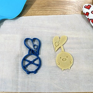 May include: Two cookie cutters on a white parchment paper surface. The cutter on the left is blue and shaped like a cartoon character with a heart on its head. The cutter on the right is yellow and shaped like a cartoon bird with a heart on its head.