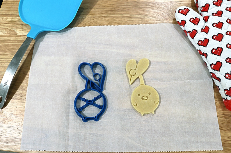 May include: Two cookie cutters on a white parchment paper surface. The cutter on the left is blue and shaped like a cartoon character with a heart on its head. The cutter on the right is yellow and shaped like a cartoon bird with a heart on its head.