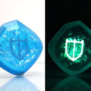 May include: A blue, square-shaped resin dice with a white shield design. The dice glows green in the dark.