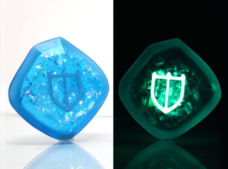May include: A blue, square-shaped resin dice with a white shield design. The dice glows green in the dark.