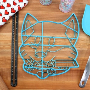 May include: A blue plastic cookie cutter shaped like a cartoon dog with a tongue sticking out. The cutter is on a wooden surface with a rolling pin, a measuring tape, and a spatula.