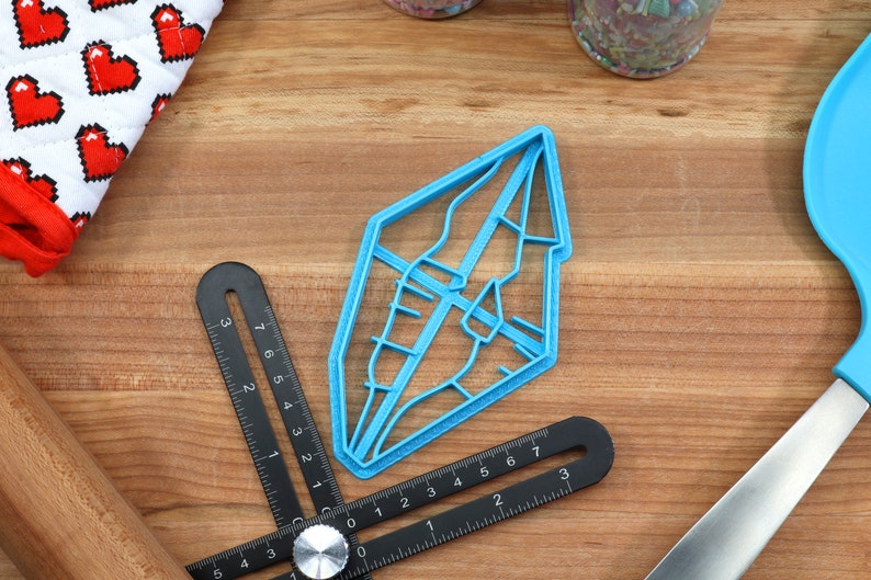 以下が含まれることがあります： ダイヤモンドの形をした青いプラスチック製のクッキーカッター。内側に詳細なデザインが施されています。カッターは、定規と麺棒のある木製の表面の上に置かれています。