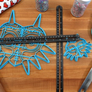 May include: Two blue plastic cookie cutters shaped like the Majora's Mask from the Legend of Zelda video game. The larger cutter is 6 inches wide and the smaller cutter is 3 inches wide. A black ruler is placed on top of the larger cutter and a black ruler is placed to the right of the smaller cutter.