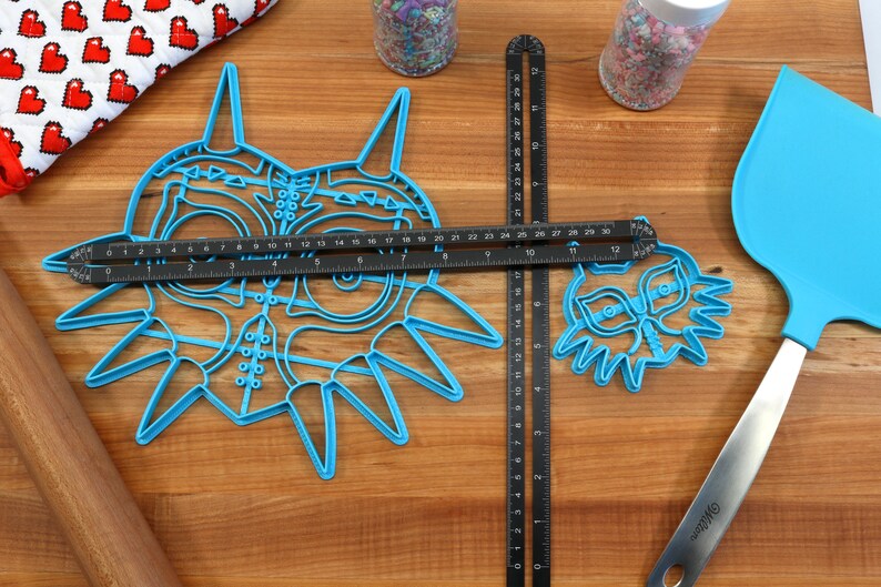 May include: Two blue plastic cookie cutters shaped like the Majora's Mask from the Legend of Zelda video game. The larger cutter is 6 inches wide and the smaller cutter is 3 inches wide. A black ruler is placed on top of the larger cutter and a black ruler is placed to the right of the smaller cutter.