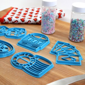 May include: A set of six blue plastic cookie cutters shaped like a roulette wheel, dice, slot machine, playing cards, and two different styles of a money bag. The cutters are on a wooden surface with two jars of sprinkles in the background.