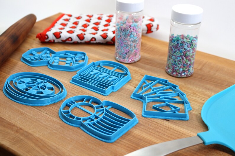 May include: A set of six blue plastic cookie cutters shaped like a roulette wheel, dice, slot machine, playing cards, and two different styles of a money bag. The cutters are on a wooden surface with two jars of sprinkles in the background.