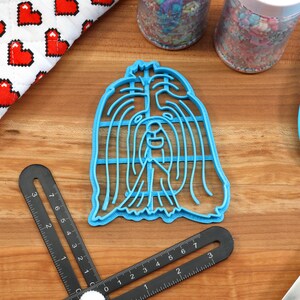 May include: Blue plastic cookie cutter shaped like a Shih Tzu dog's head. The cutter is on a wooden surface with a ruler and a blue spatula.