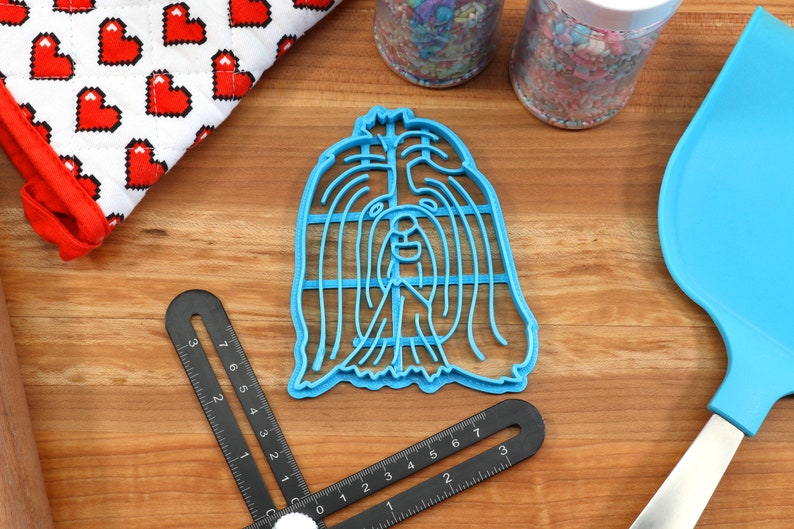 May include: Blue plastic cookie cutter shaped like a Shih Tzu dog's head. The cutter is on a wooden surface with a ruler and a blue spatula.