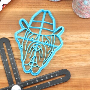 Peut inclure: Un emporte-pi&egrave;ce en plastique bleu en forme de bouledogue portant un chapeau. L'emporte-pi&egrave;ce est sur une surface en bois.