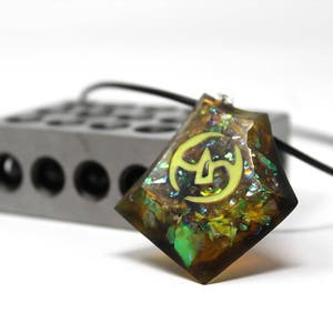 May include: A green, brown, and gold iridescent resin pendant with a yellow symbol inside. The pendant is on a black cord.