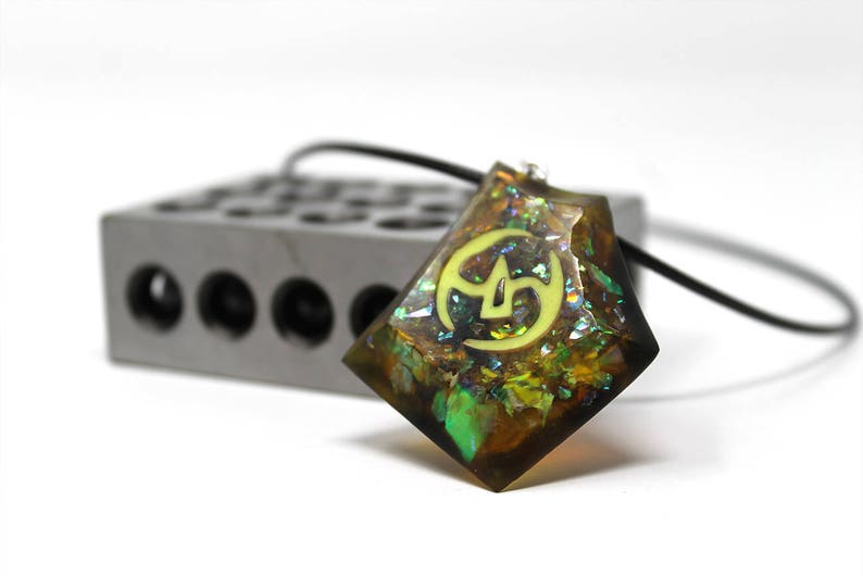 May include: A green, brown, and gold iridescent resin pendant with a yellow symbol inside. The pendant is on a black cord.