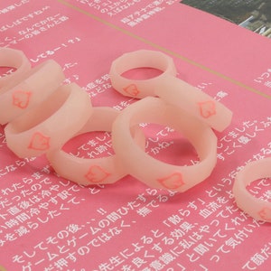 May include: A close-up of several translucent pink silicone rings with a heart design. The rings are arranged on a pink surface.