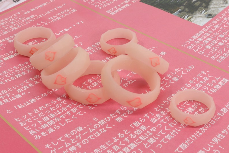 May include: A close-up of several translucent pink silicone rings with a heart design. The rings are arranged on a pink surface.