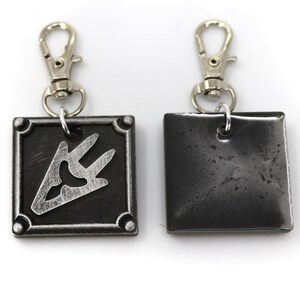 May include: A silver metal keychain with a black square charm featuring a silver symbol. The back of the charm is plain black.