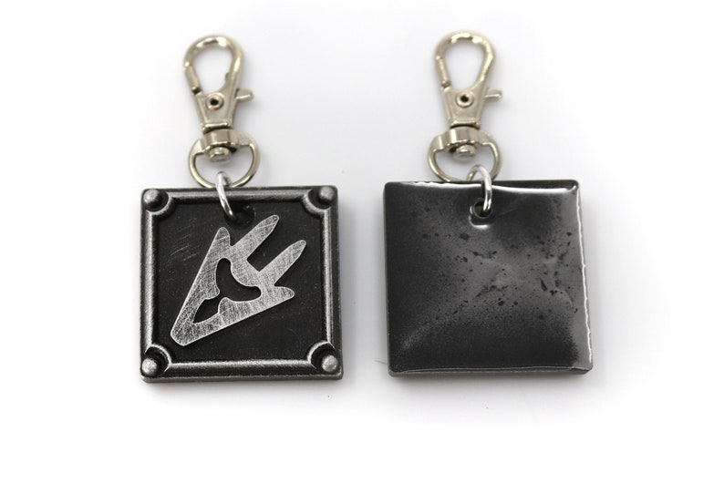 May include: A silver metal keychain with a black square charm featuring a silver symbol. The back of the charm is plain black.
