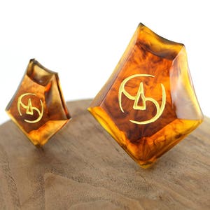May include: Two amber-colored dice with gold-colored markings. The dice are shaped like a diamond and have a swirl pattern. The dice are sitting on a wooden surface.