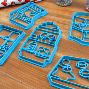 May include: Five blue plastic cookie cutters shaped like different buildings. The cutters are on a wooden surface.