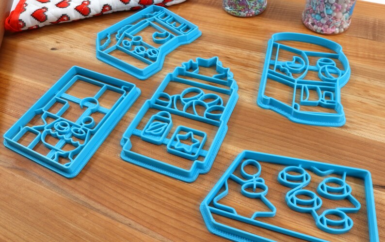 May include: Five blue plastic cookie cutters shaped like different buildings. The cutters are on a wooden surface.