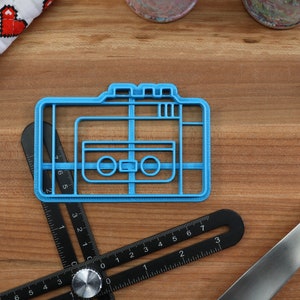 May include: A blue plastic cookie cutter shaped like a vintage cassette tape. The cookie cutter is on a wooden surface.