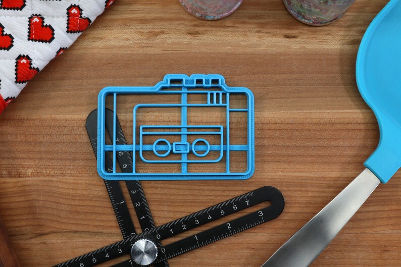 May include: A blue plastic cookie cutter shaped like a vintage cassette tape. The cookie cutter is on a wooden surface.
