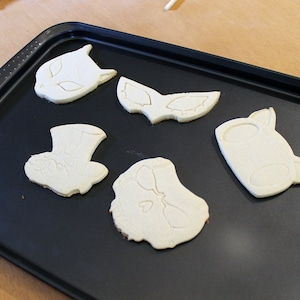 May include: Five unbaked sugar cookies on a black baking sheet. The cookies are shaped like a cat, a mask, a hat, and two other shapes.