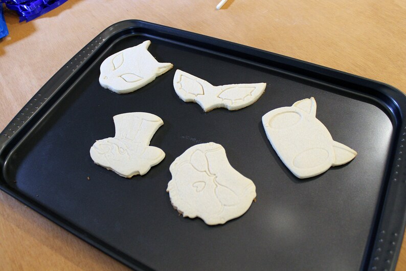 May include: Five unbaked sugar cookies on a black baking sheet. The cookies are shaped like a cat, a mask, a hat, and two other shapes.