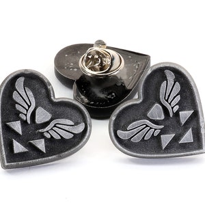 May include: Three silver heart-shaped pins with a winged symbol and a triangular design. The pins are on a white background.