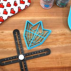 May include: A blue plastic geometric cookie cutter in the shape of an arrow on a wooden surface with a black ruler and a blue spatula.
